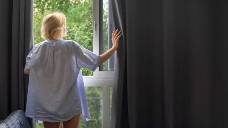 mulher abrindo estilosa cortina blackout para olhar a janela. Confira modelos de cortinas blackout